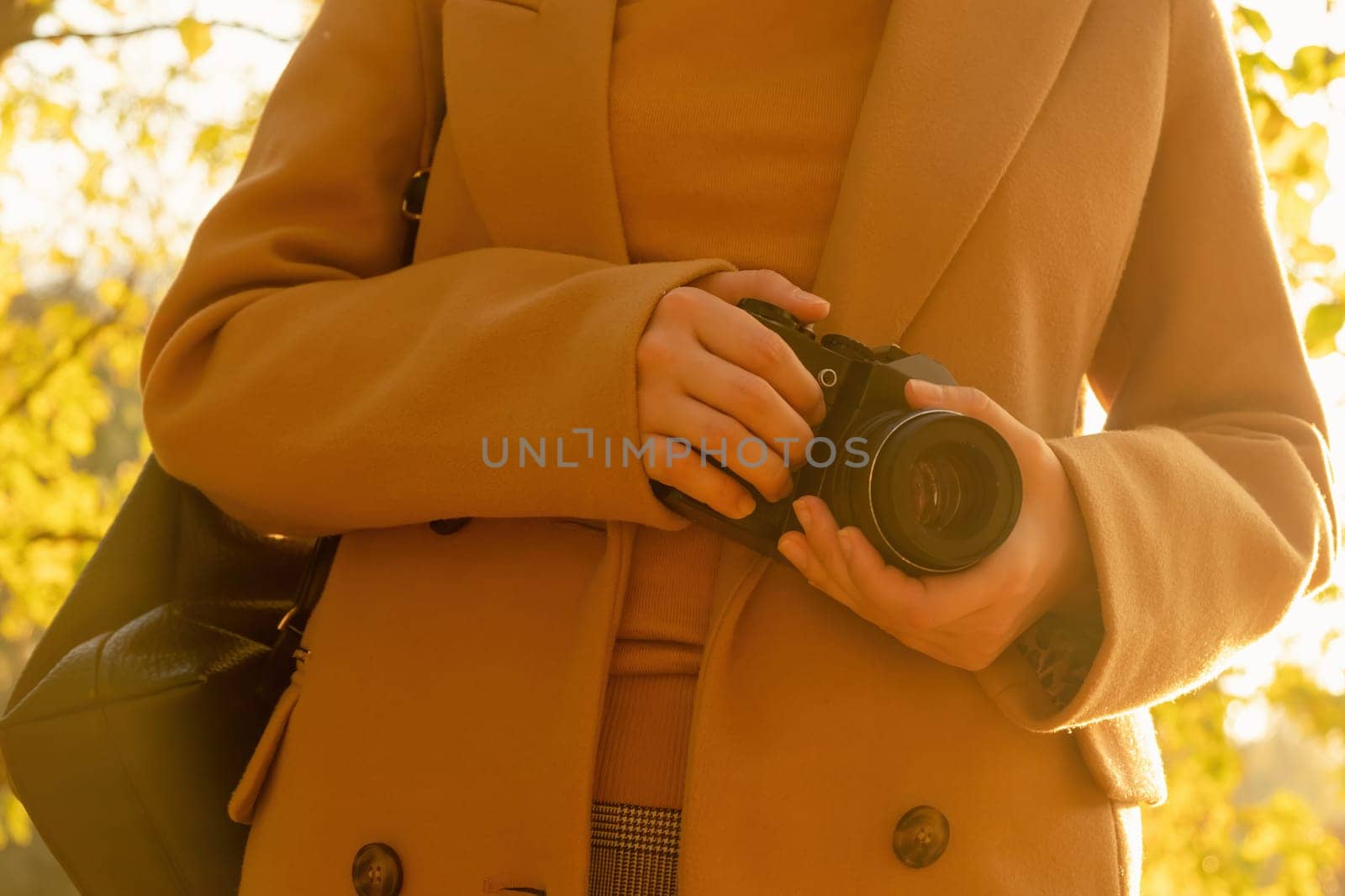 A woman in a brown coat holding a retro camera in her hands against trees with yellow leaves in the park