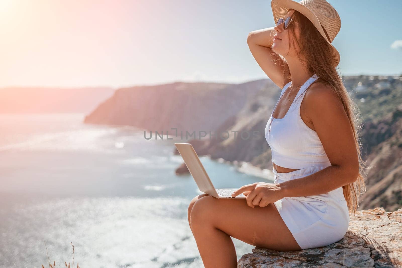 Successful business woman in yellow hat working on laptop by the sea. Pretty lady typing on computer at summer day outdoors. Freelance, travel and holidays concept.