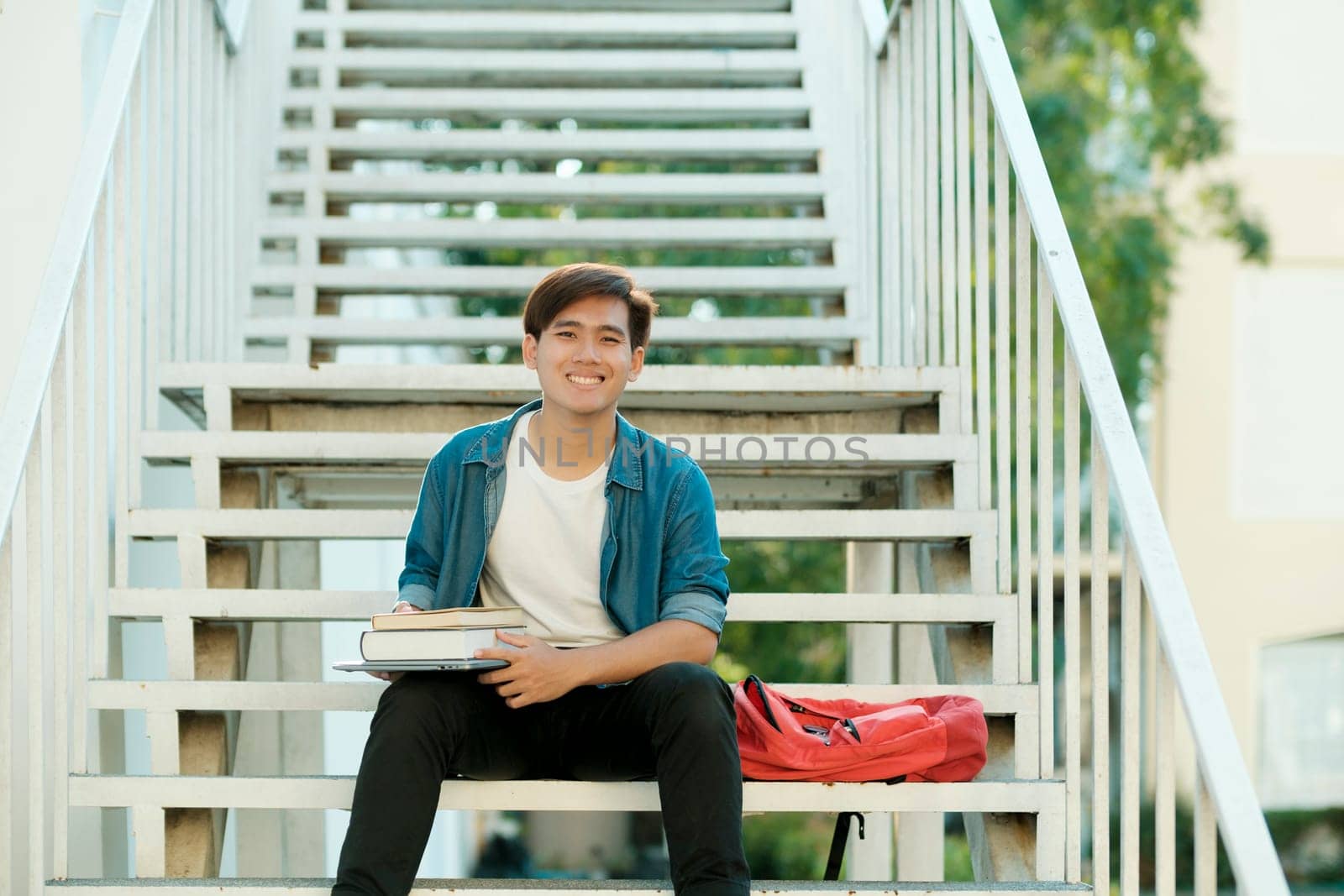 Young male college student in casual clothings looking at camera, holding books, and sitting outdoor on stairs to study and read books preparing for school project and research. Education concept.