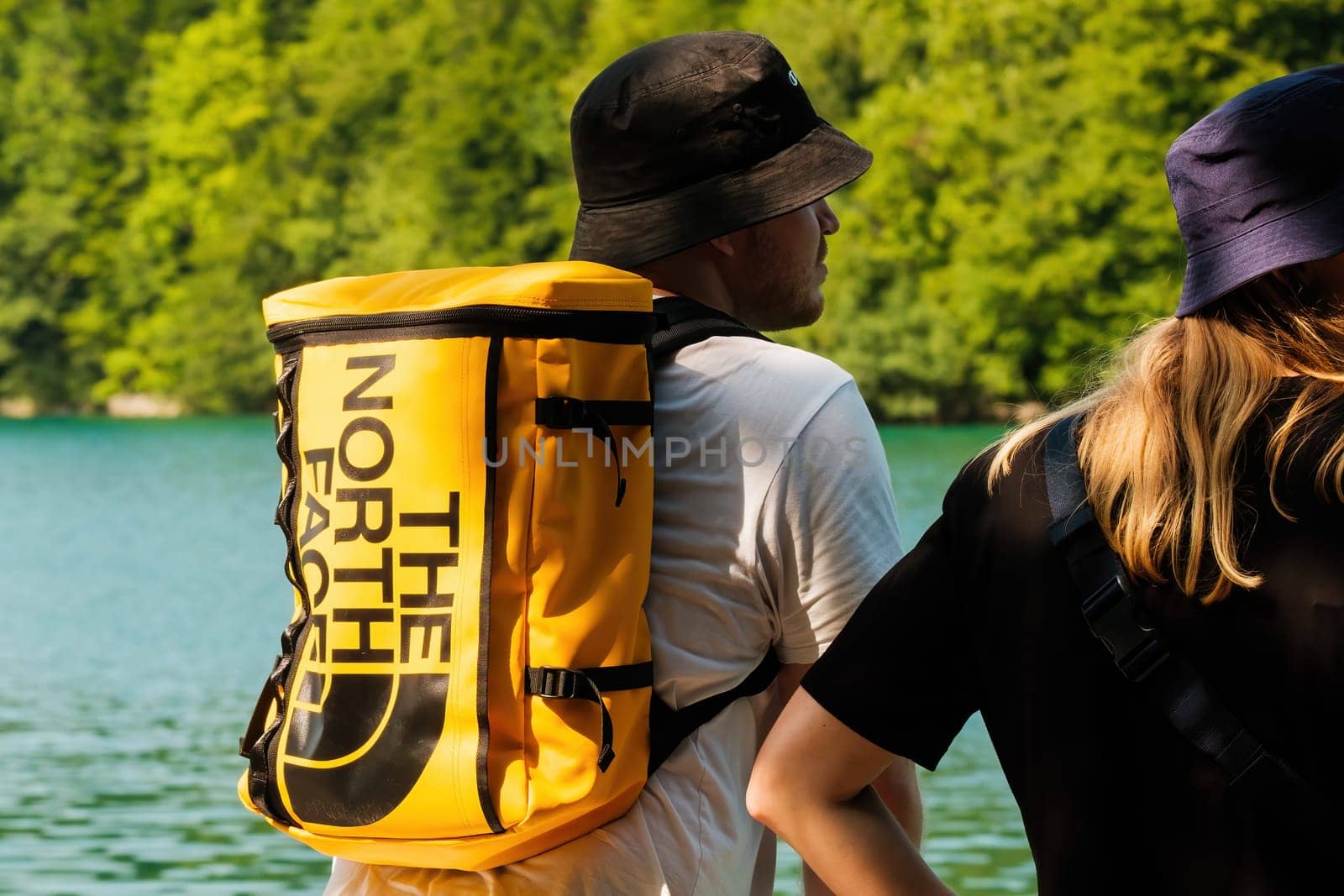 RIJEKA, CROATIA - JULY 10 2022: Tourists wearing dark panamas walk along blue lake surrounded by trees. Man with yellow backpack The North Face and woman on excursion looking at lake back view