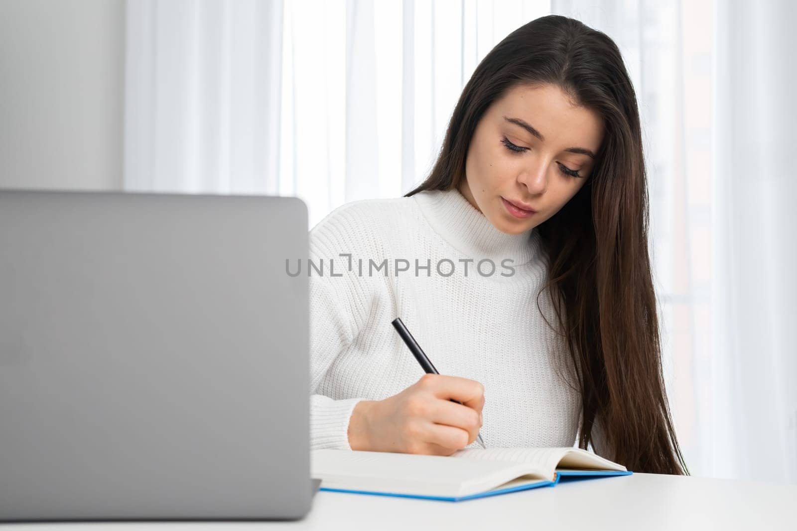 Young woman takes online courses via notebook making notes while listening to teacher. Attentive student sits at table in room studying online at home