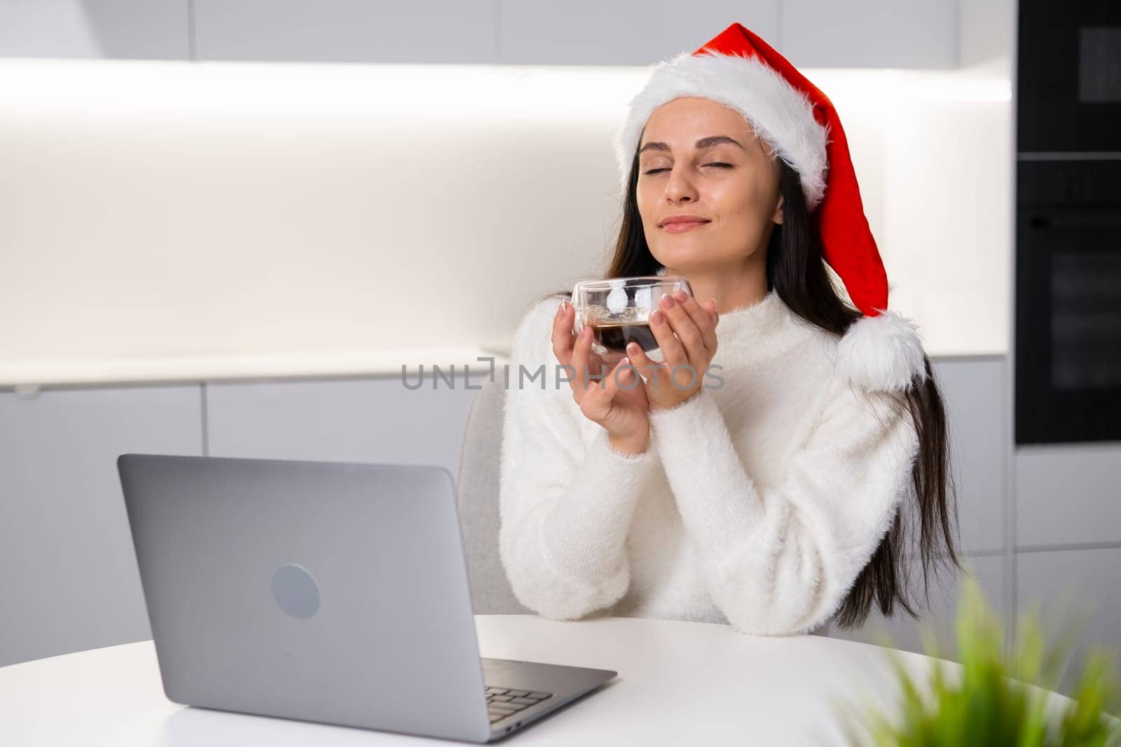 A dreamy woman in a Santa Claus hat closes her eyes and enjoys her coffee on a coffee break while working at home using her laptop