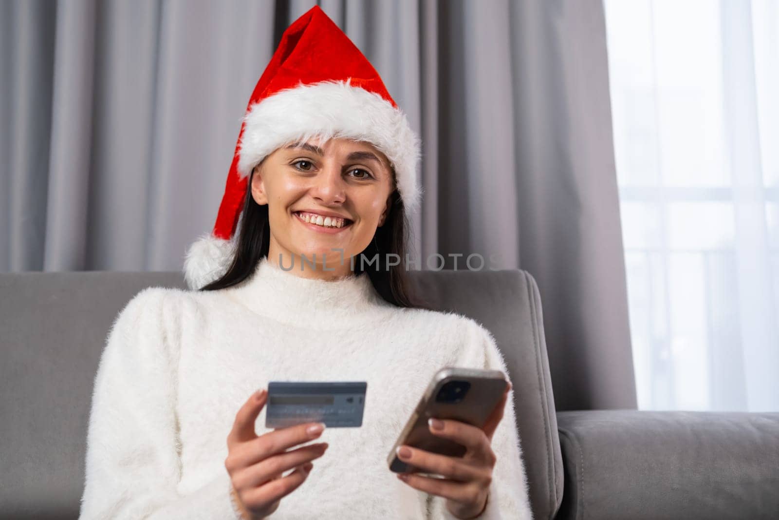 Portrait of a smiling beautiful woman wearing a Santa Claus hat holding a credit card and smartphone for online shopping. Preparation for Christmas and New Year party.