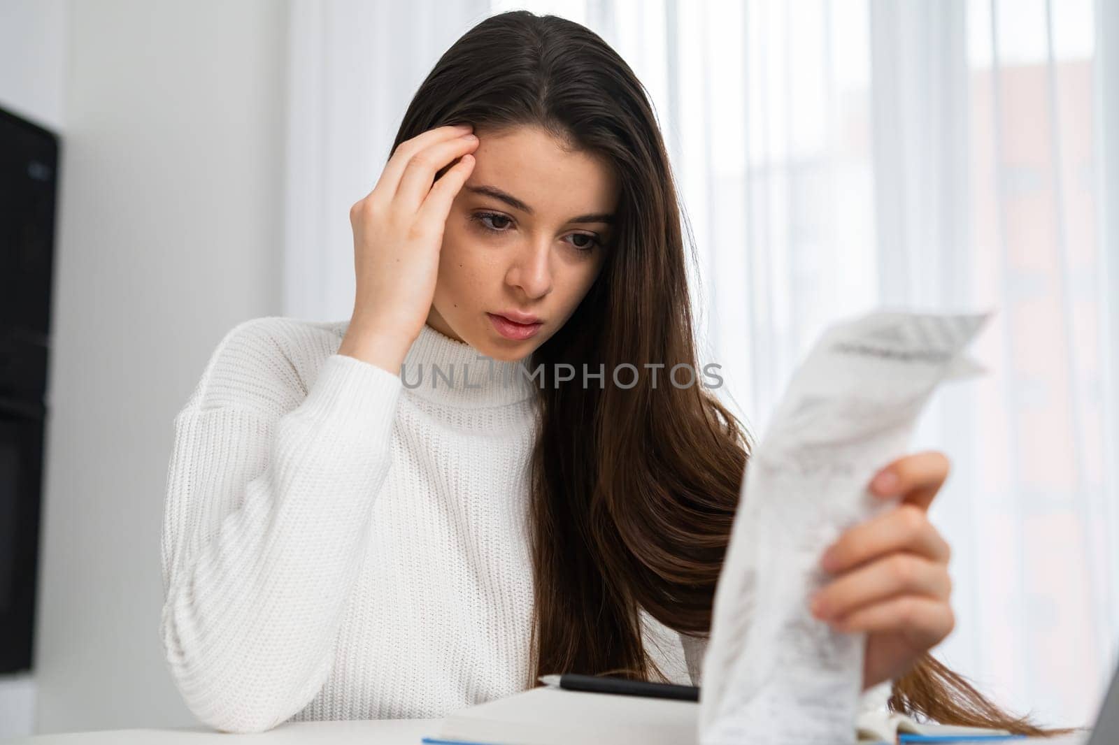 Upset woman looks at spent amount in check after shopping. Young freelancer sits at table near Notebook checking list of purchases at home