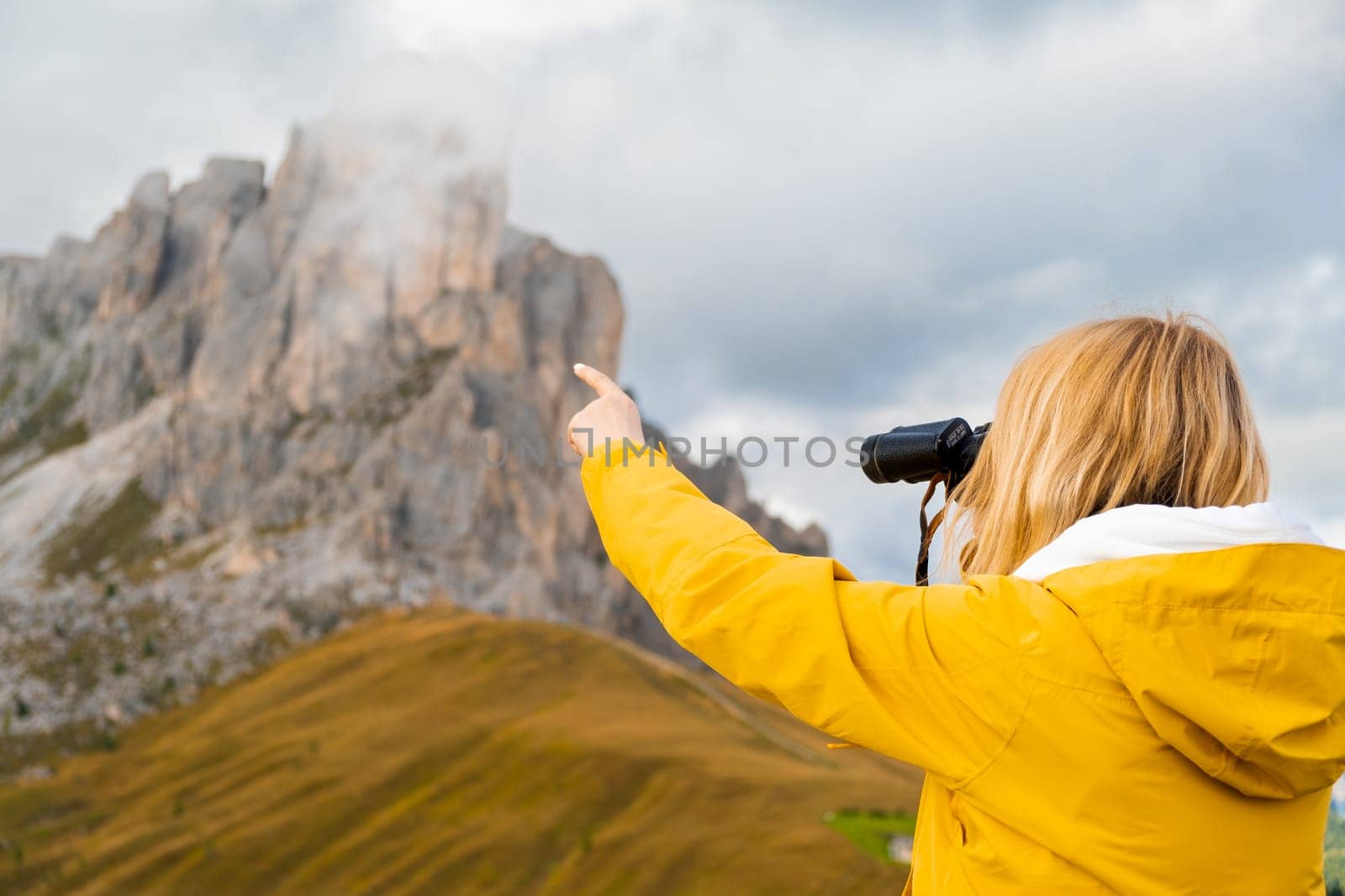 Blonde woman looks at Passo Giau pass through binoculars pointing finger on rocky mountain covered with clouds. Female tourist enjoys hiking in Italian Alps