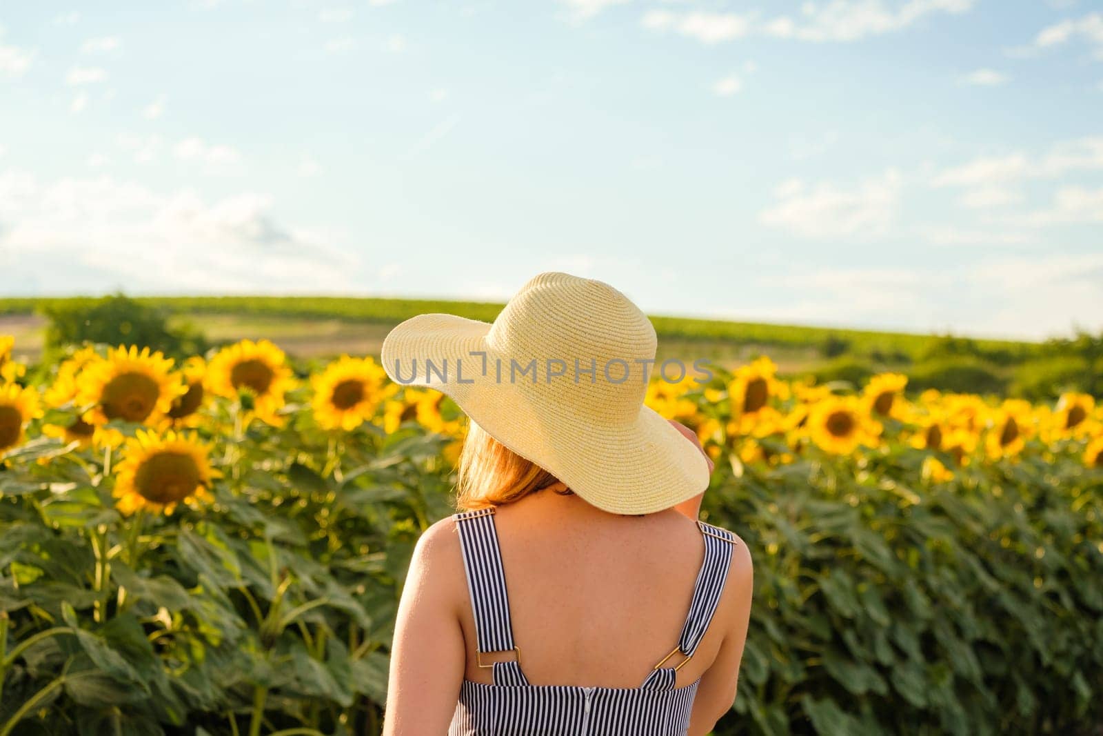 Young woman in wicker straw hat stands against sunflower field on sunny summer day. Lady enjoys nature in countryside backside close view