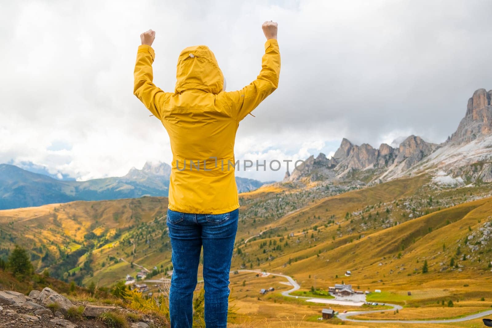 A young woman in the hood and yellow jacket enjoying the mountains view of the Italian Dolomites and raise your hands. Travel concept