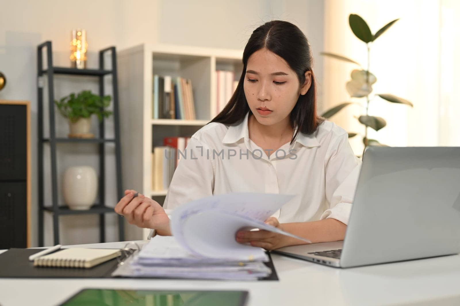 Concentrated businesswoman siting in front of laptop computer at working desk and checking marketing reports.