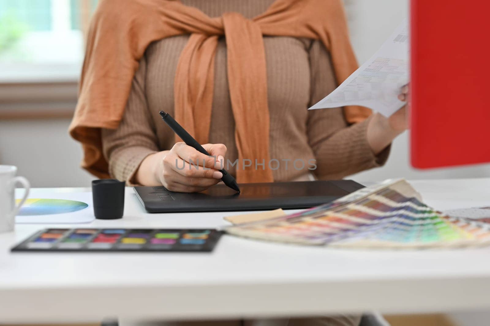 Young creative woman using graphic tablet and working with color swatch samples at workstation.