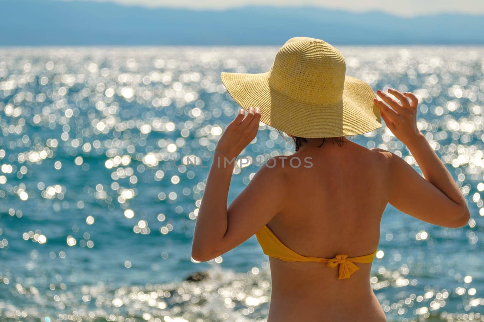 Woman in yellow bikini and straw hat looks at blue sea on warm day. Lady holds headdress with hands against water sparkling in sun close back view