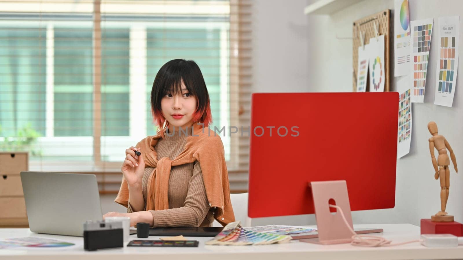 Portrait of young asian woman designer sitting at graphic studio in front of computer monitor and looking confidently to camera by prathanchorruangsak