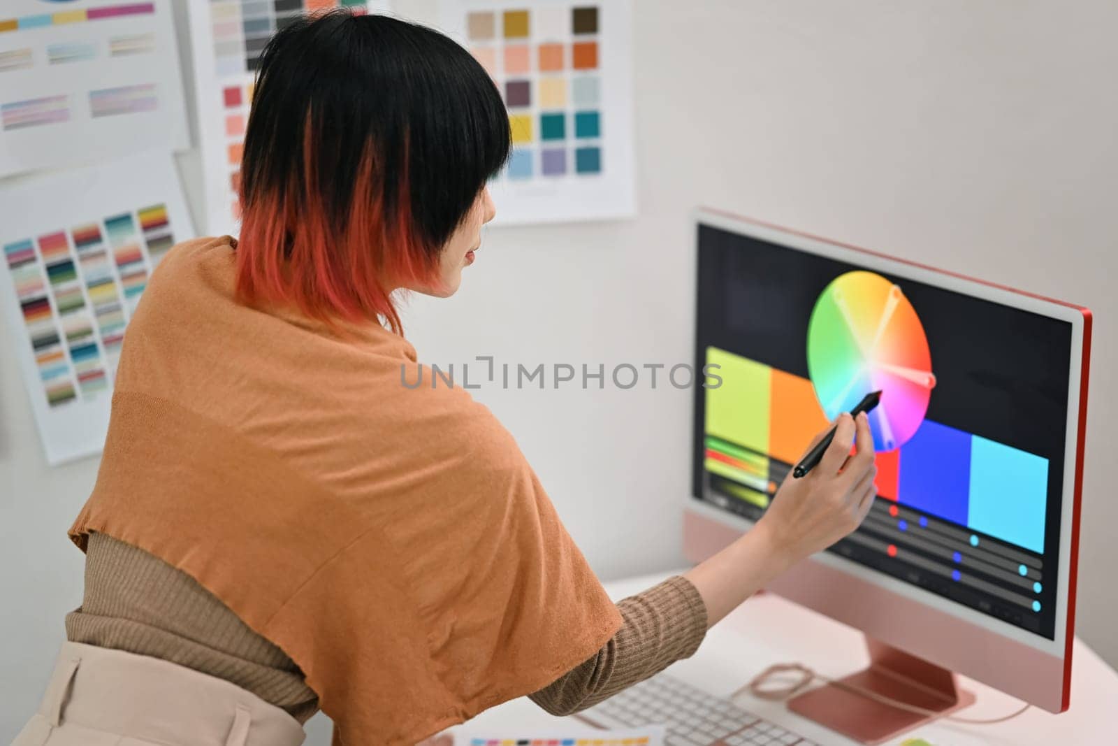 Side view of asian female graphic designer choosing colors from color swatch samples on computer display in creative office.
