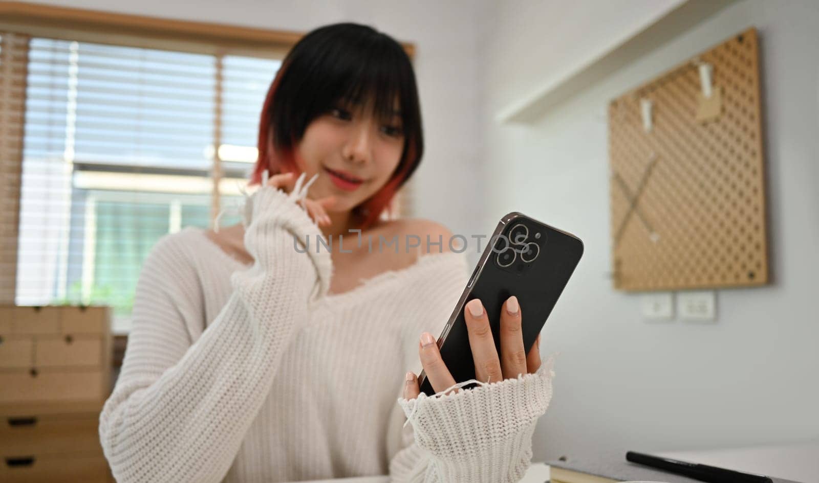 Smiling young asian woman texting messages, chatting online on mobile phone, sitting in modern home office.