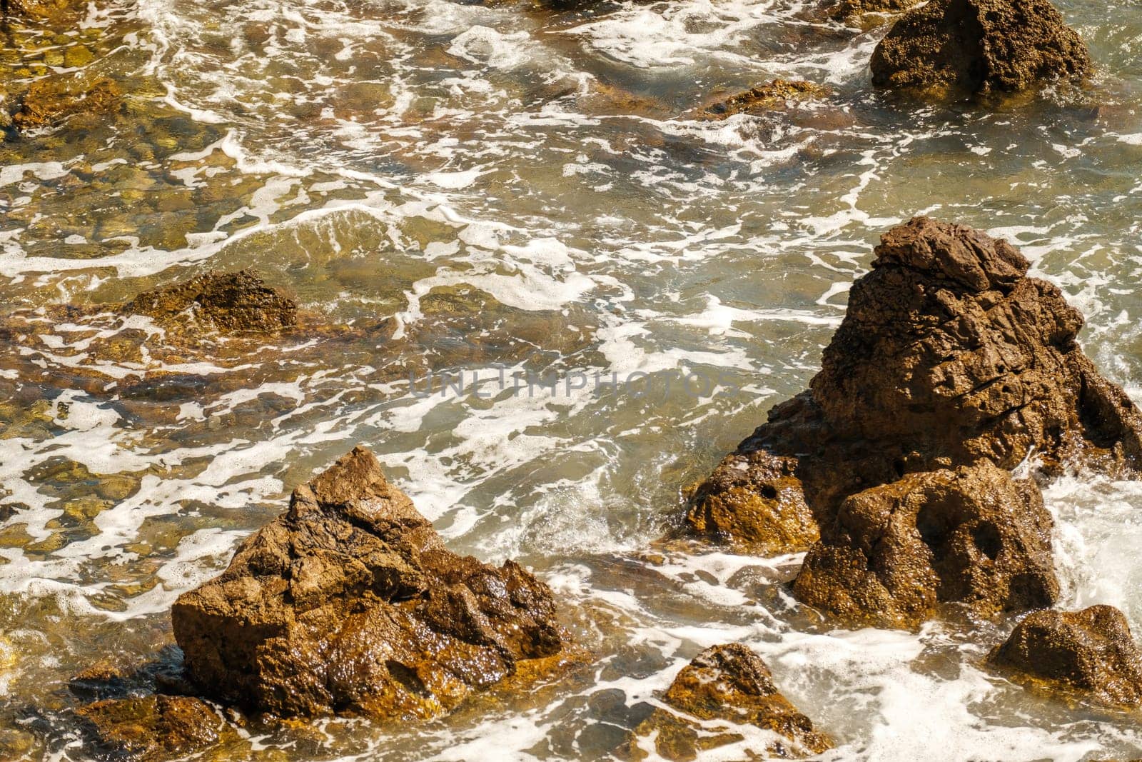 Small ocean waves crashing of rocks on shallow coastline at high tide at bright sunlight. Transparent waves create foams rolling on stones