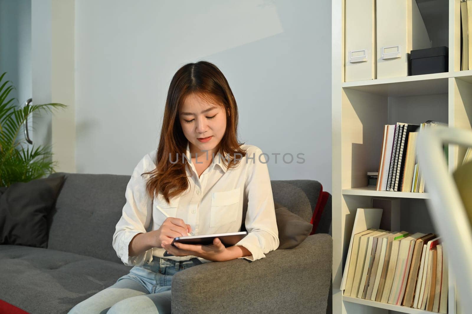 Young female freelancer in white shirt using digital tablet on couch at home. Communication, technology concept.