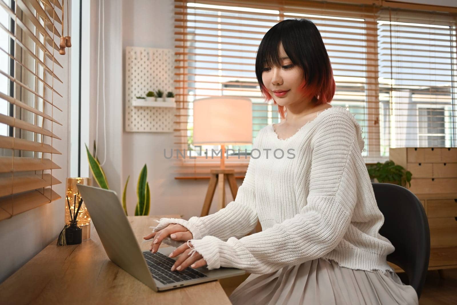 Pretty young Asian woman using laptop browsing internet and checking videos on social networks, sitting in a cozy living room by prathanchorruangsak