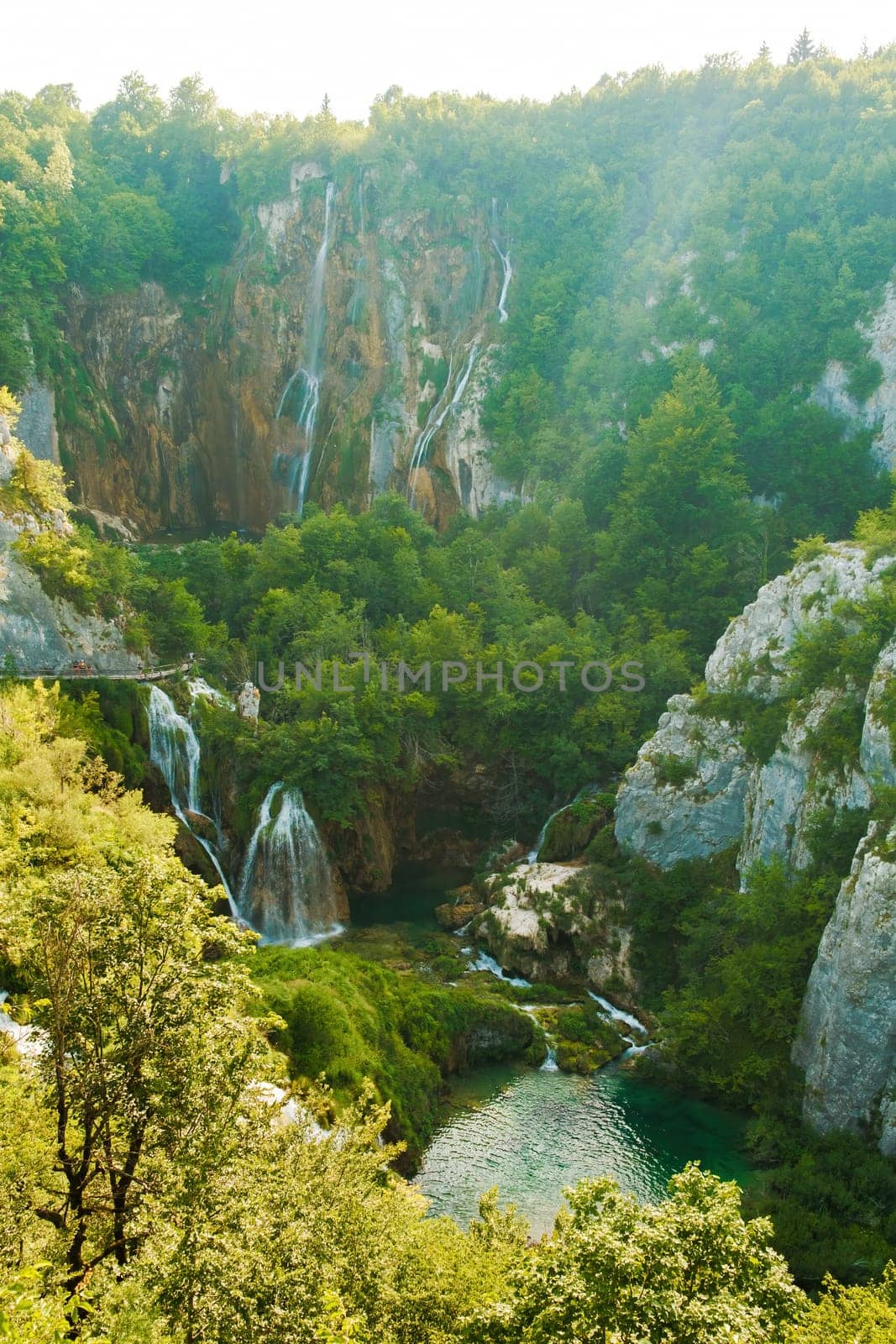 Waterfalls with cascades falling from steep rocks into canyon. Forestry and bare cliffs surround gorge with clear blue lake on bottom of Plitvice lakes upper view