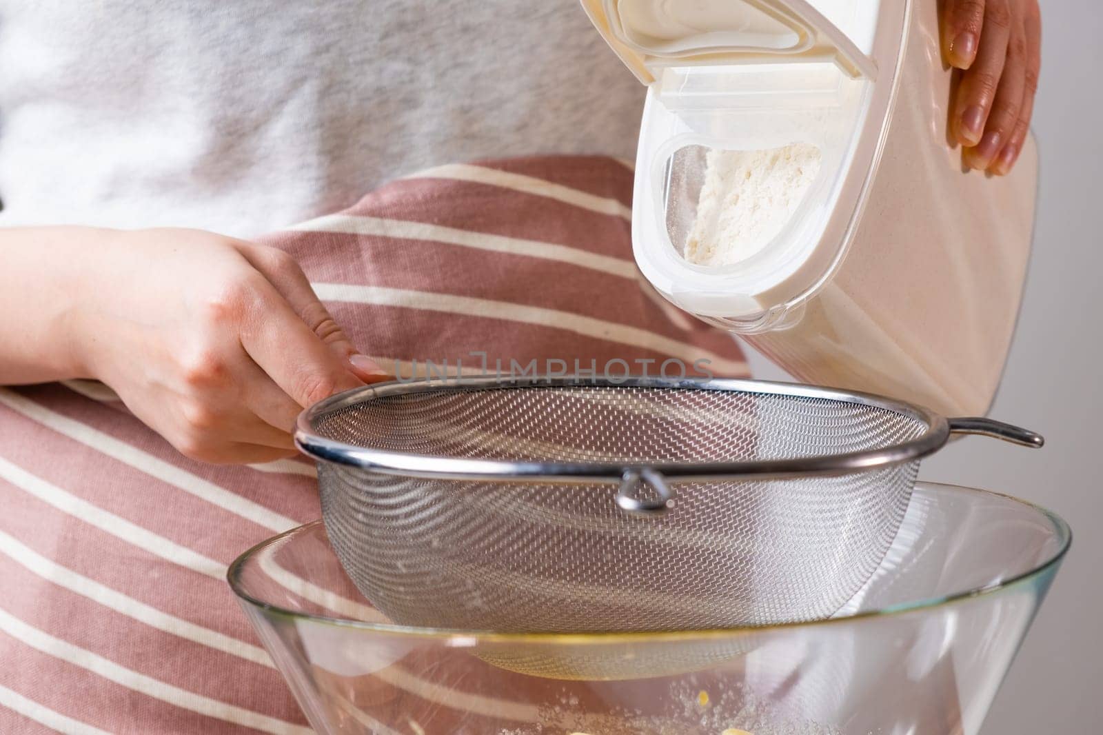 Close up woman sifts wheat flour through a sieve into mixing bowl for dough preparation for cake and bakery on the black background. Knead the dough for homemade pastry.