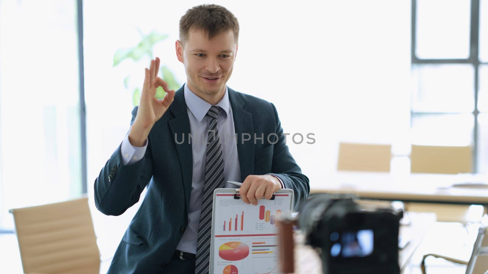 Successful businessman coach with paperwork with graphs in hands showing ok gesture in front of camera in office. Business blogging concept