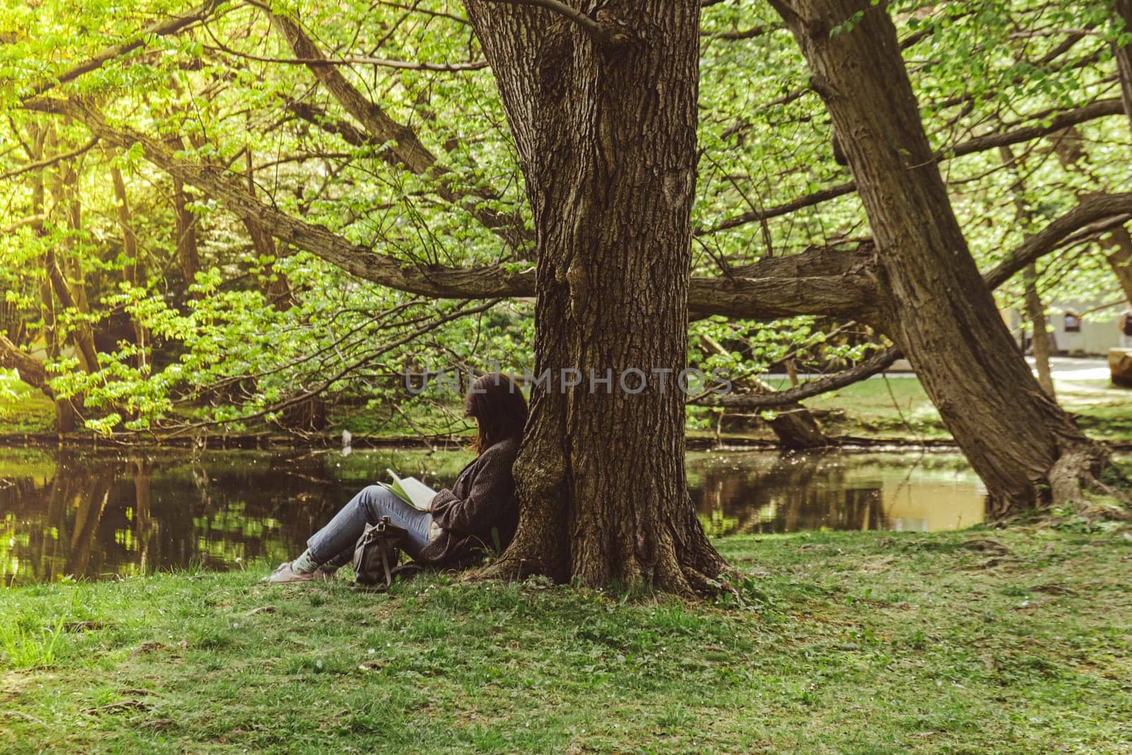 Attractive young woman reading book while sitting on grass in green public park. Springtime outdoors. Greenery unity with nature. Spend free time on open air. Education concept