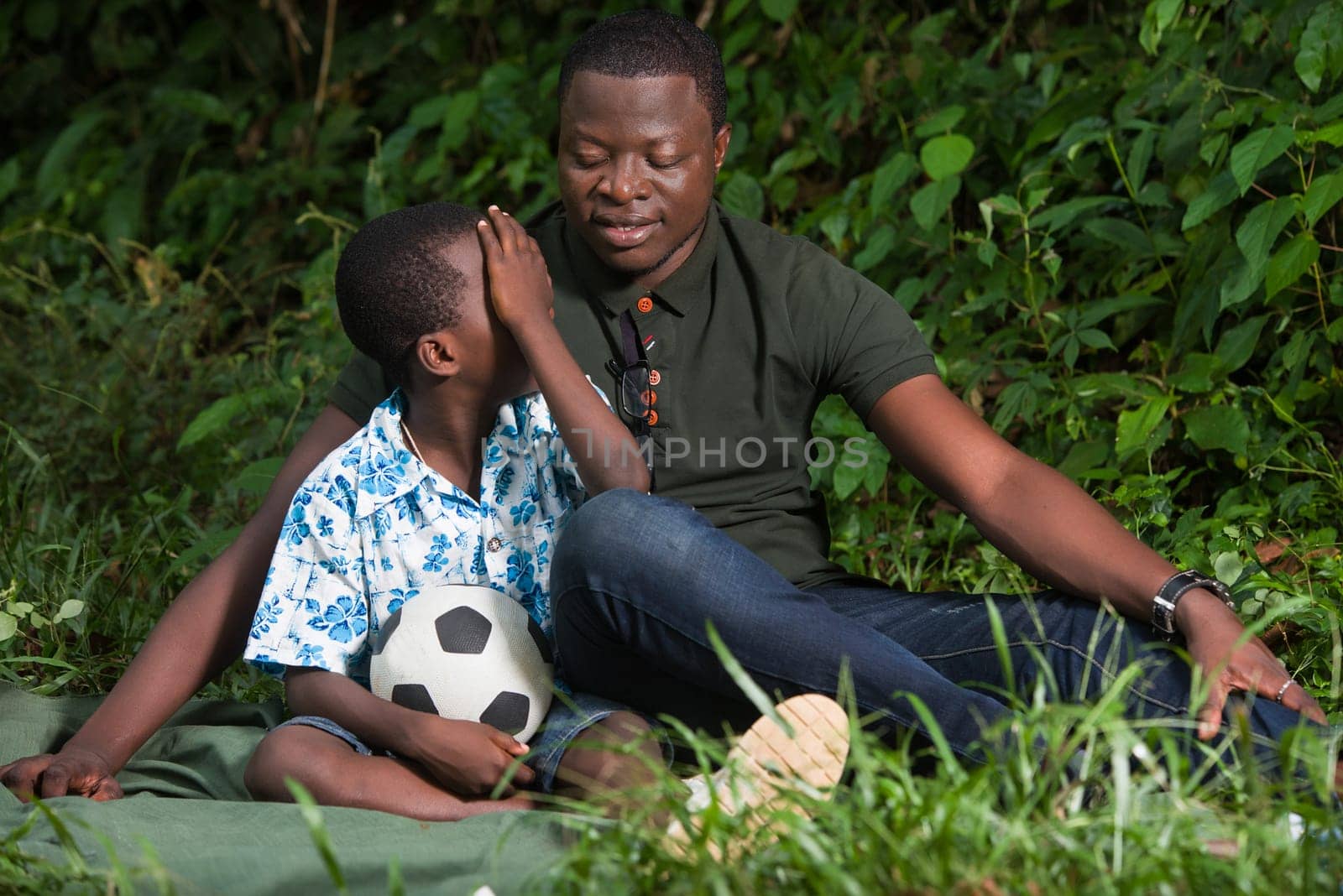 a young man sitting in a park watching his son with his hand on his face smiling.