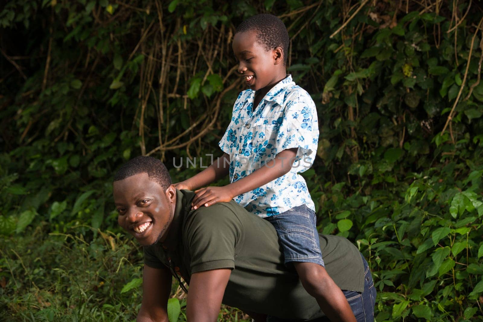 a young man on all fours in a park with his son on his back smiling at the camera.