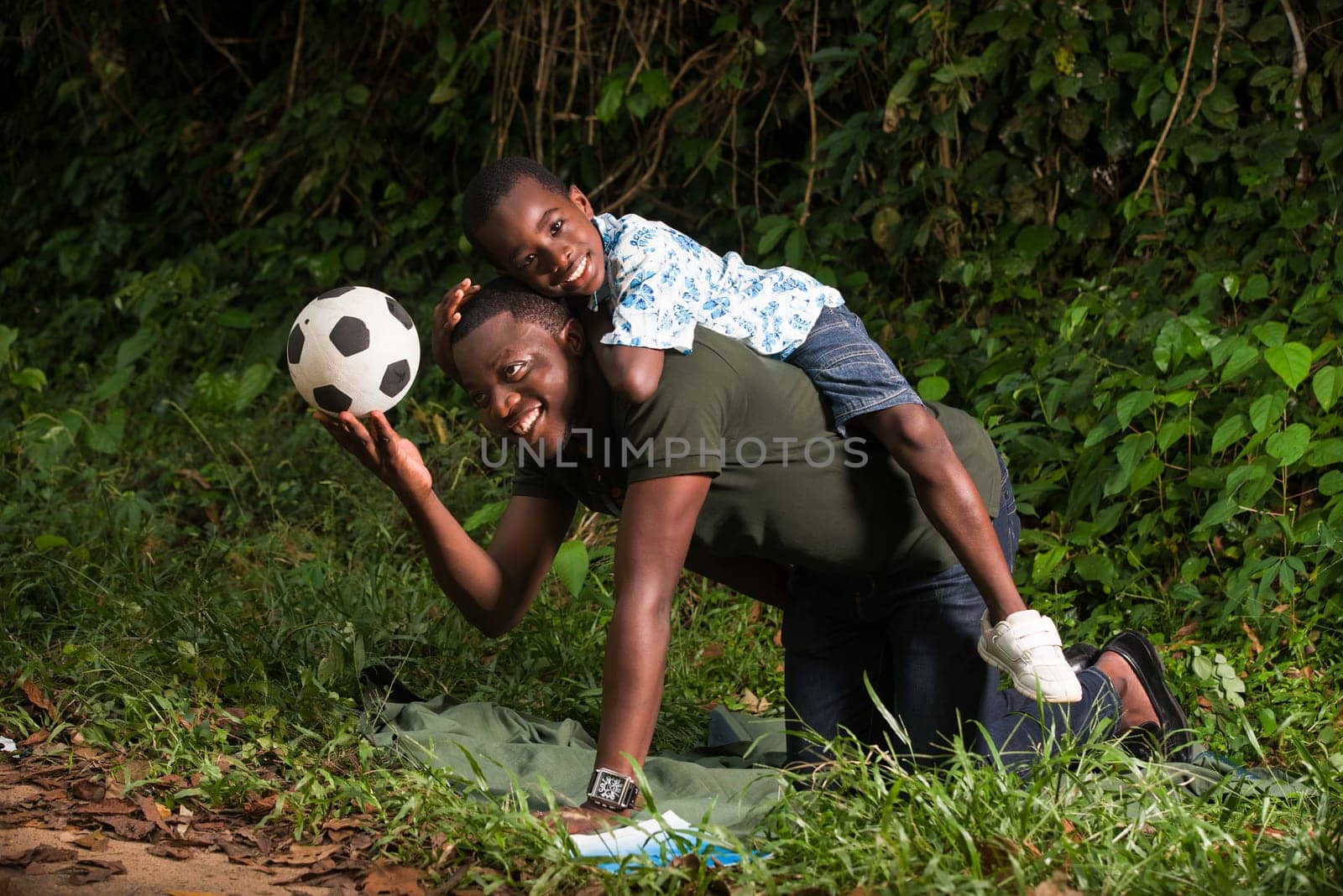 a young man on all fours in a park with his son on his back smiling at the camera.