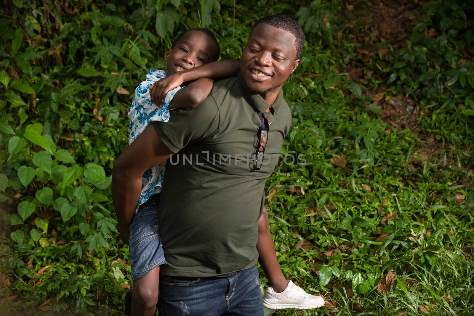 a young man kneeling in a park with his child on his back smiling.