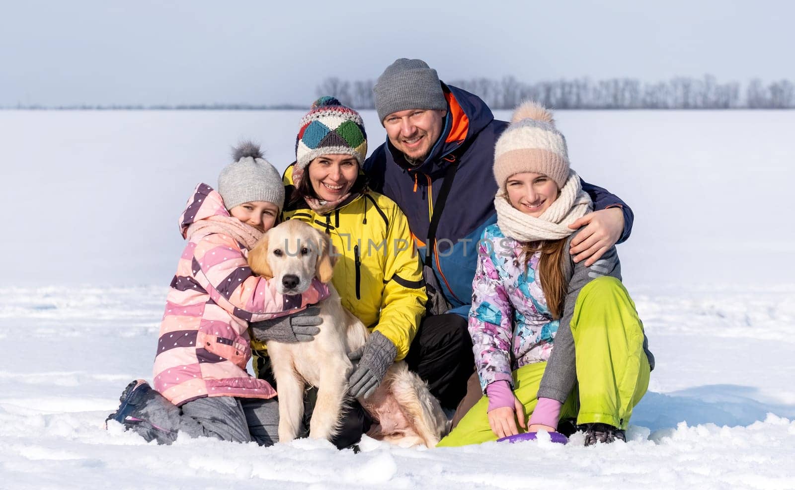 Happy family with golden retriever dog sitting on snow in winter time and smiling. Mother, father and daughters with doggy pet outdoors in Christmas season