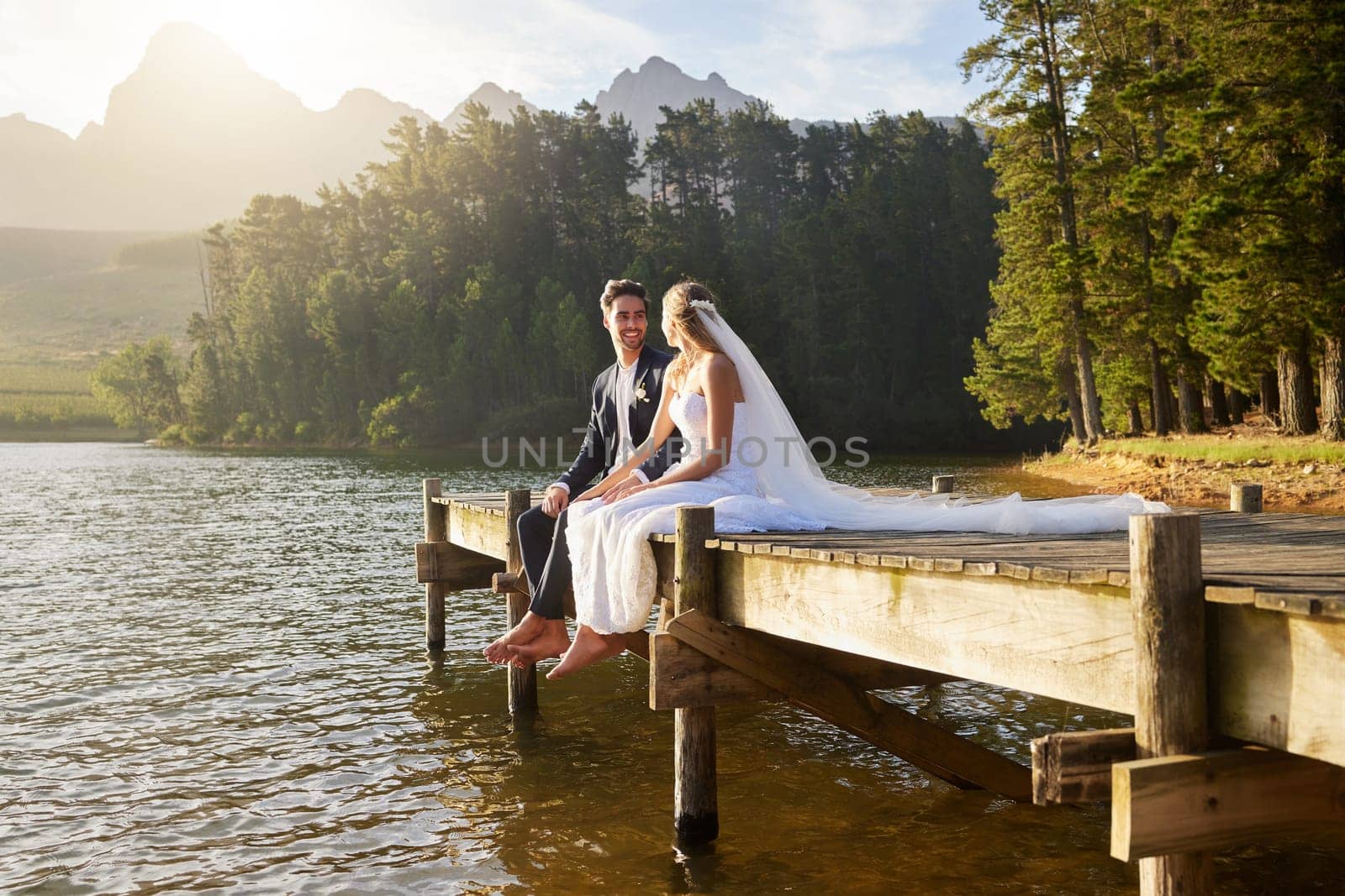 Forest, lake and a married couple on a pier in celebration together after a wedding ceremony of tradition. Marriage, love or romance with a bride and groom sitting outdoor while bonding in nature.