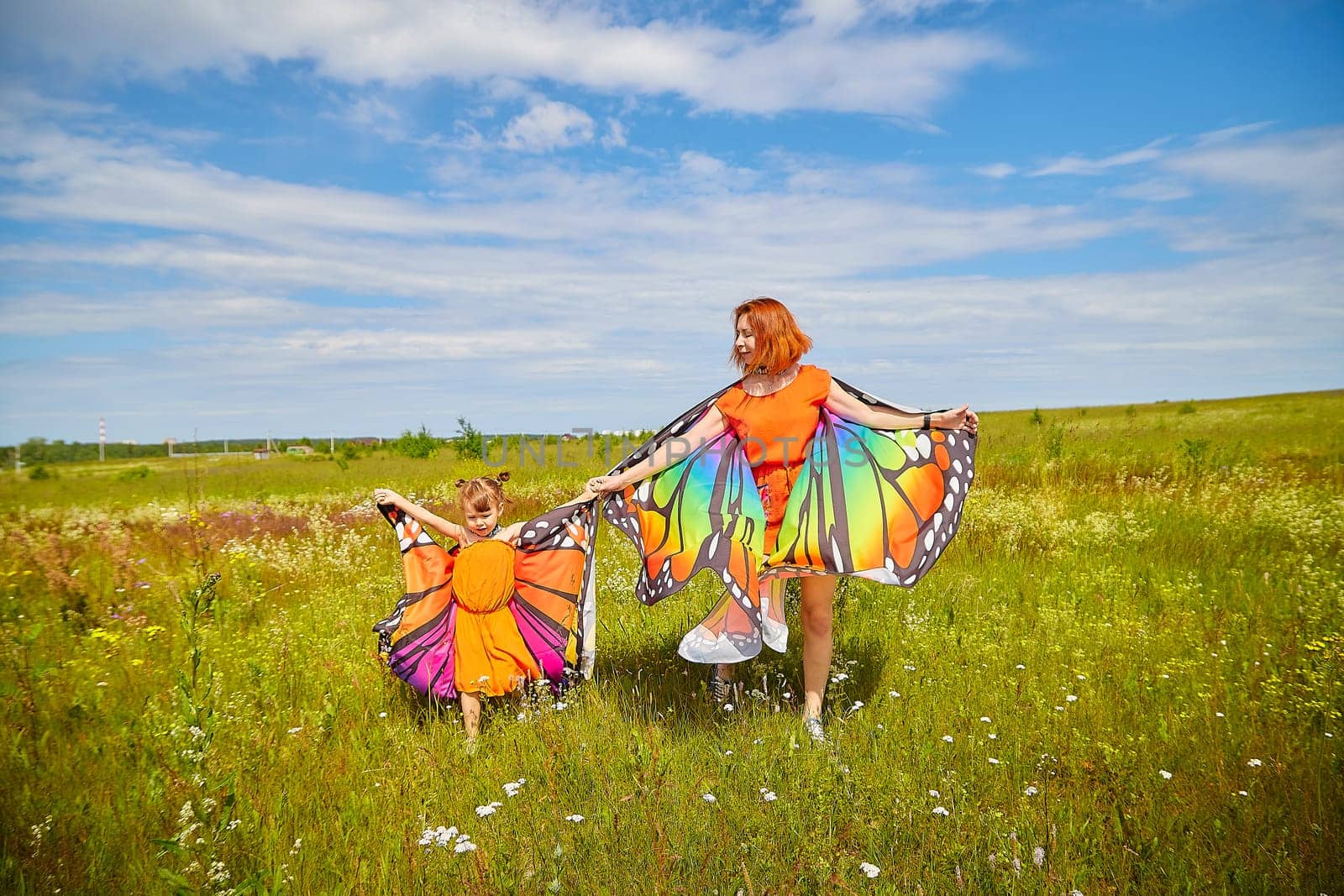 Happy female family with red haired mother and daughter with bright butterfly wings having fun on green and yellow meadow full of grass and flower in sunny summer day. Concept family love by keleny