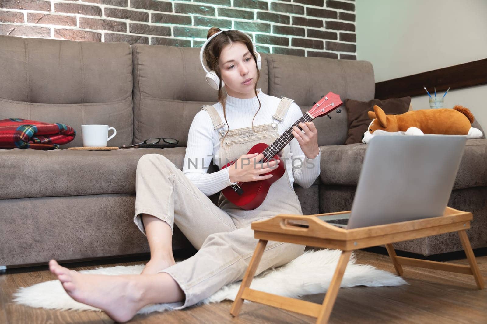 Hobbies and leisure activities during quarantine. Online training, online classes. A young woman watches a video lesson on playing the guitar, she sits on a cozy plaid with a guitar.
