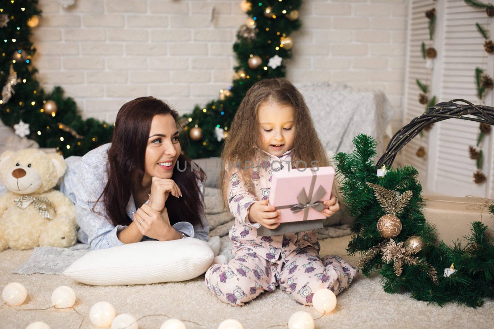 Pretty smiling child girl with mother opening magical box with Christmas gift at home