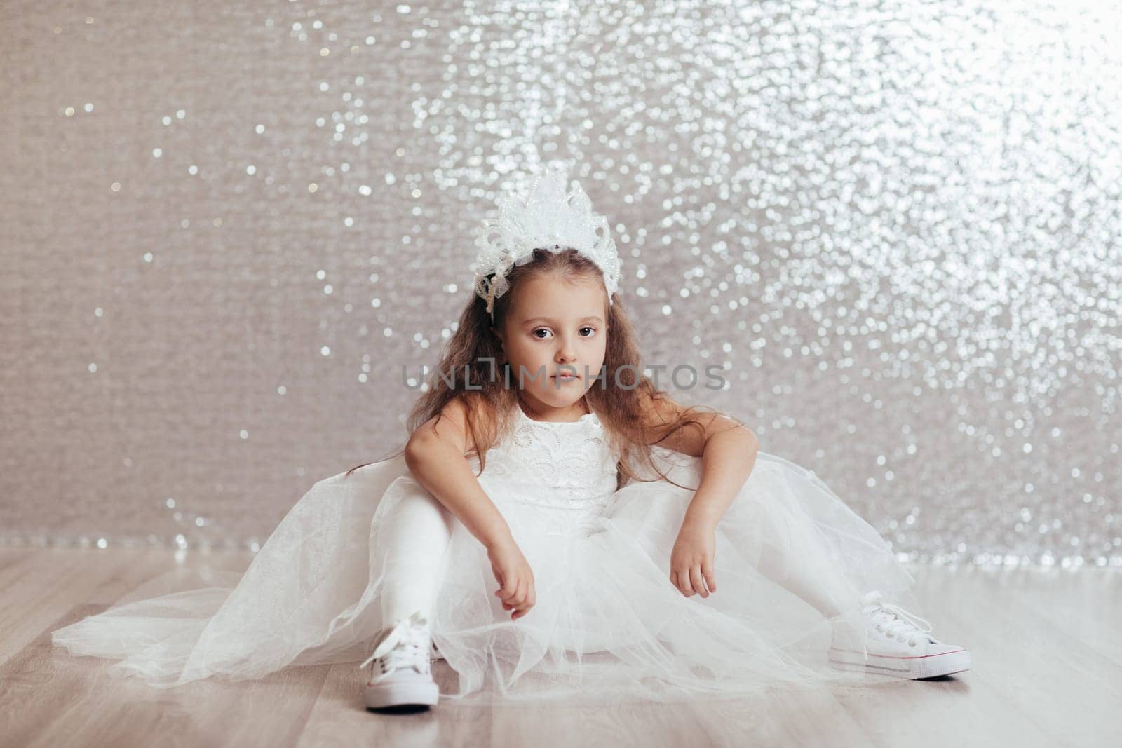 sad little child girl in blue dress sitting on the floor with confetti on background with silver bokeh. after birtday party