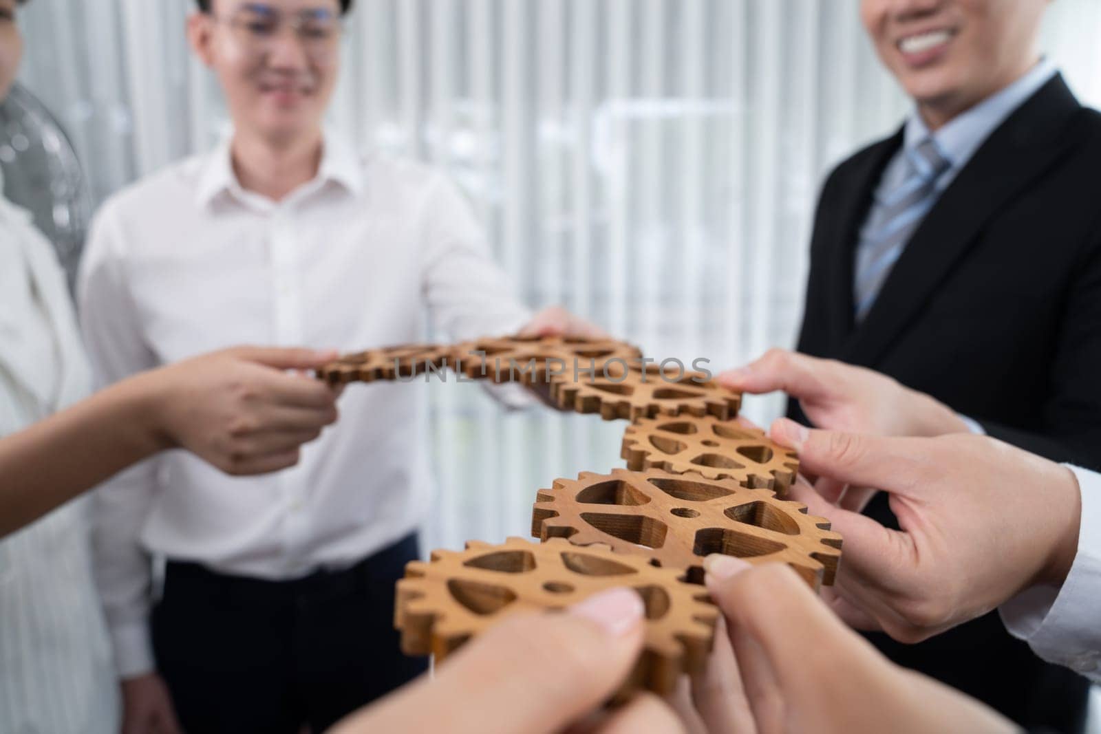 Closeup hand holding wooden gear by businesspeople wearing suit for harmony synergy in office workplace concept. Group of people hand making chain of gears into collective form for unity symbol.