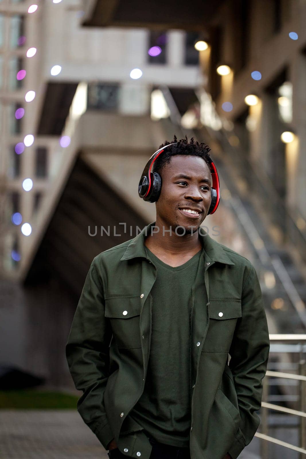 smiling african-american man in stylish jacket in wireless headphones listening music on the street of the evening city