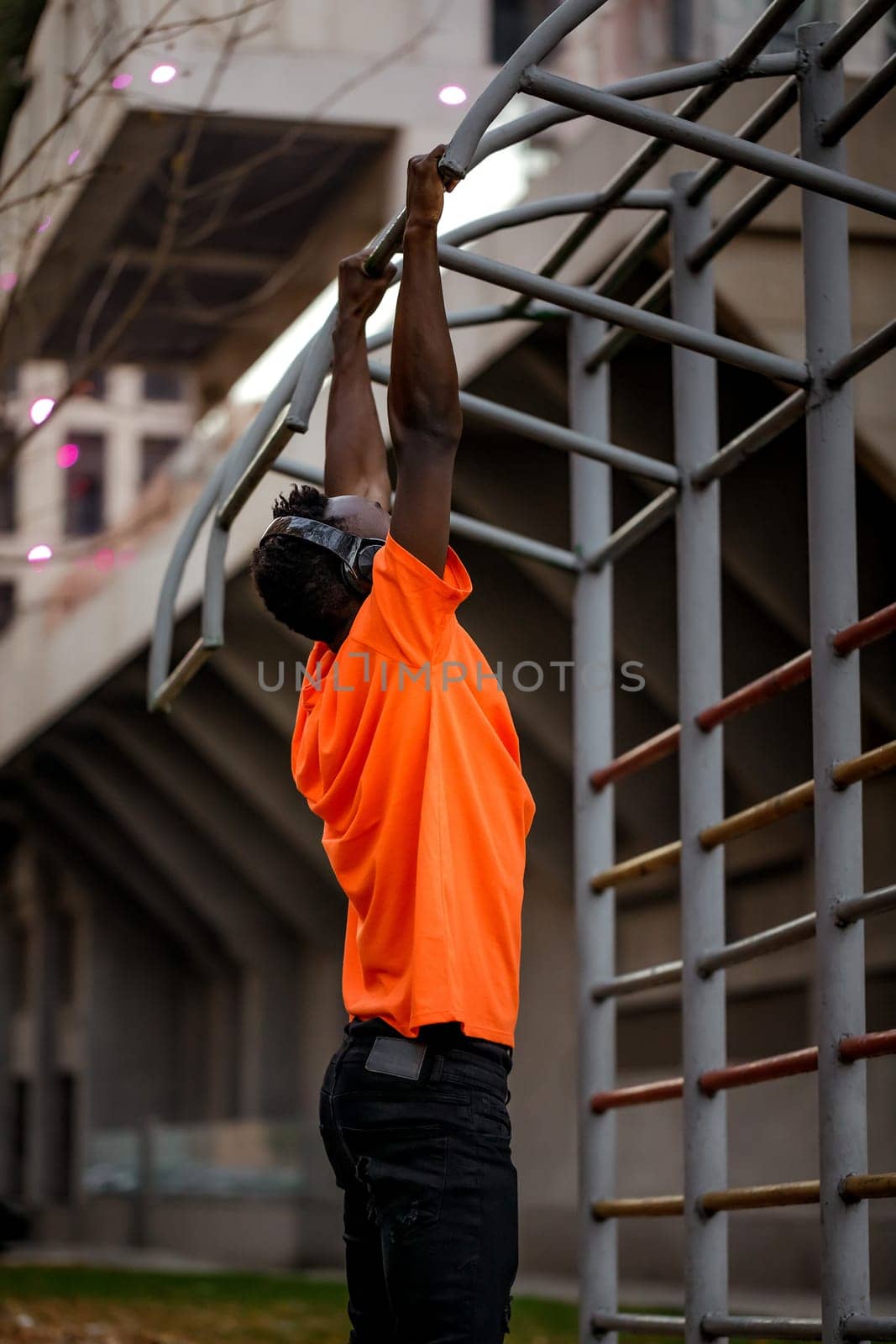 african-american man in orange t-hirt in wireless headphones pulling himself up on horizontal bars outdoor in the evening.