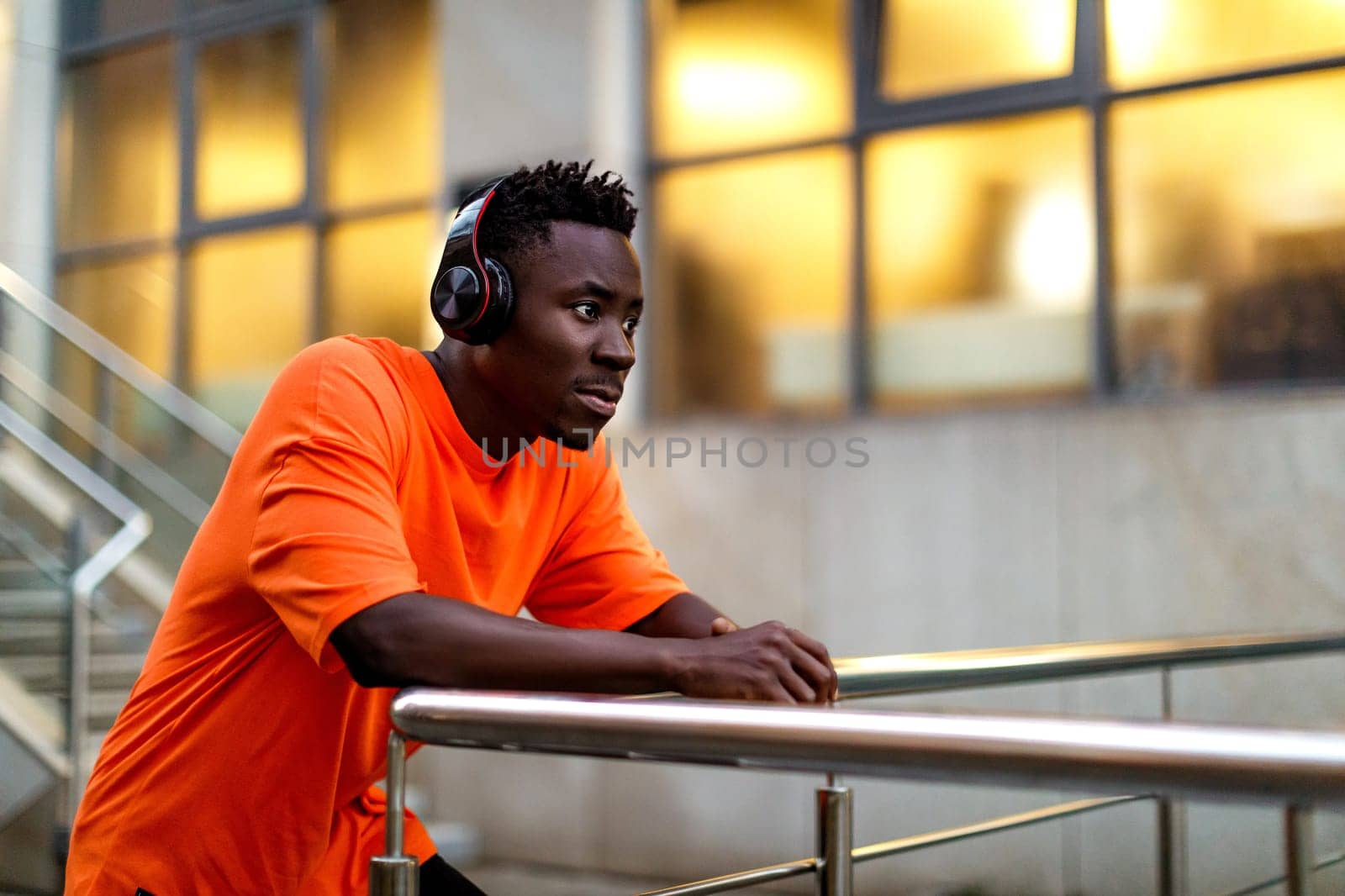african-american man in stylish orange hoodie sweatshirt in wireless headphones listening music outdoor in the evening. copy space
