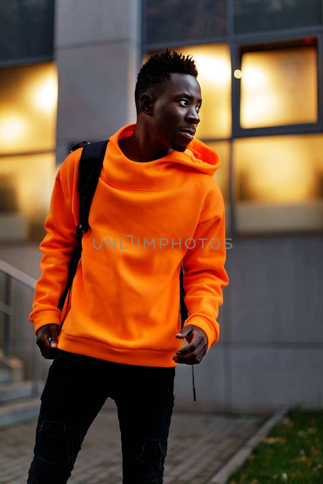 african-american man in stylish orange hoodie sweatshirt with backpack on city street in the evening
