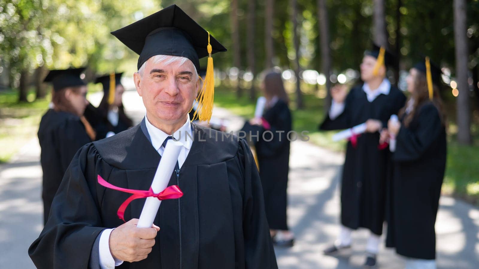 A group of graduates in robes outdoors. An elderly student rejoices at receiving a diploma