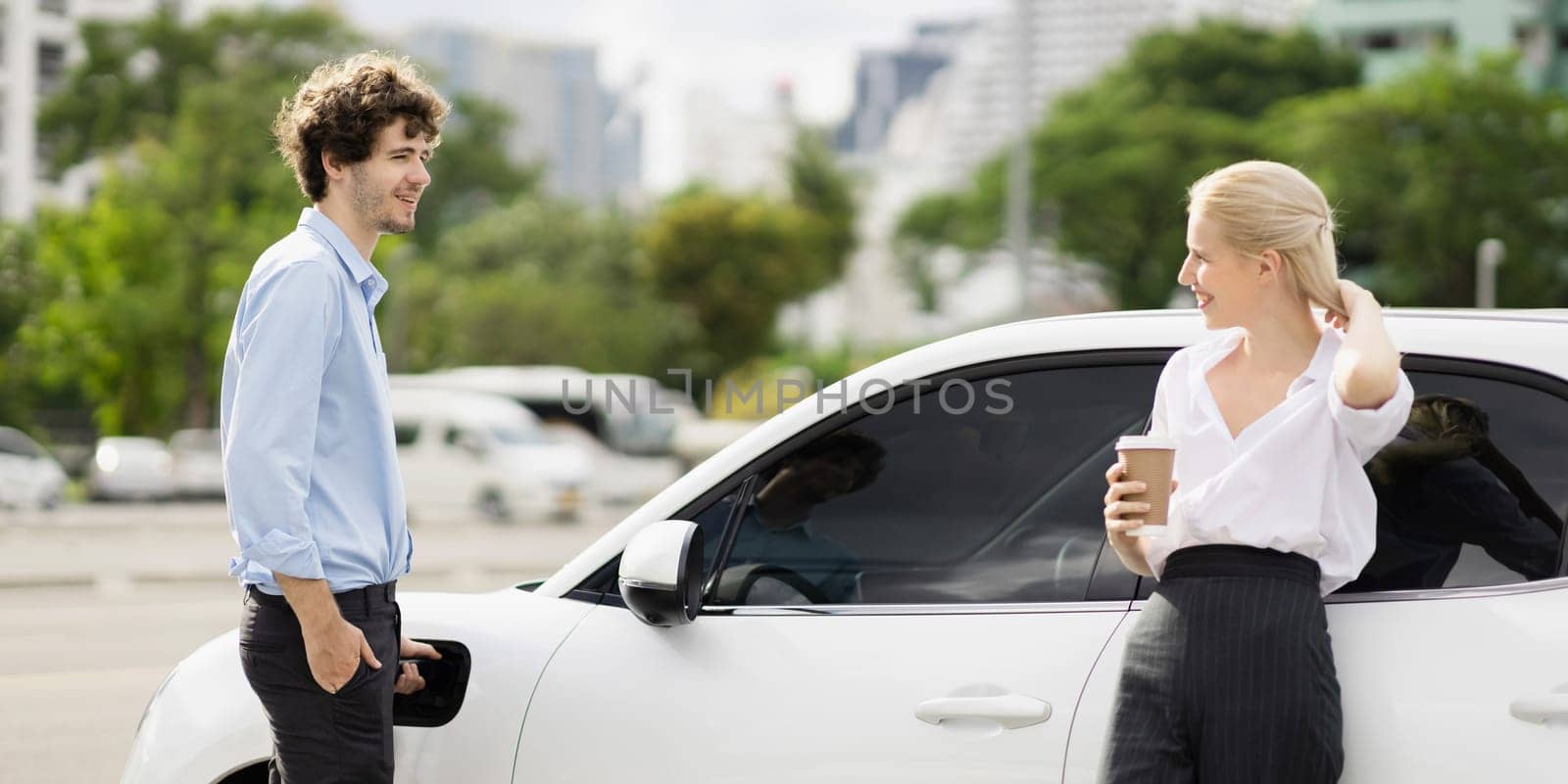 Progressive businessman and businesswoman with coffee, standing at electric car connected to charging station before driving around city center. Eco friendly rechargeable car powered by clean energy.