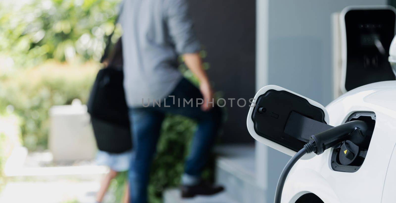 Focus electric car recharging at home charging station with blurred father and daughter walking in background. Progressive green and clean energy vehicle for healthy environment lifestyle concept.