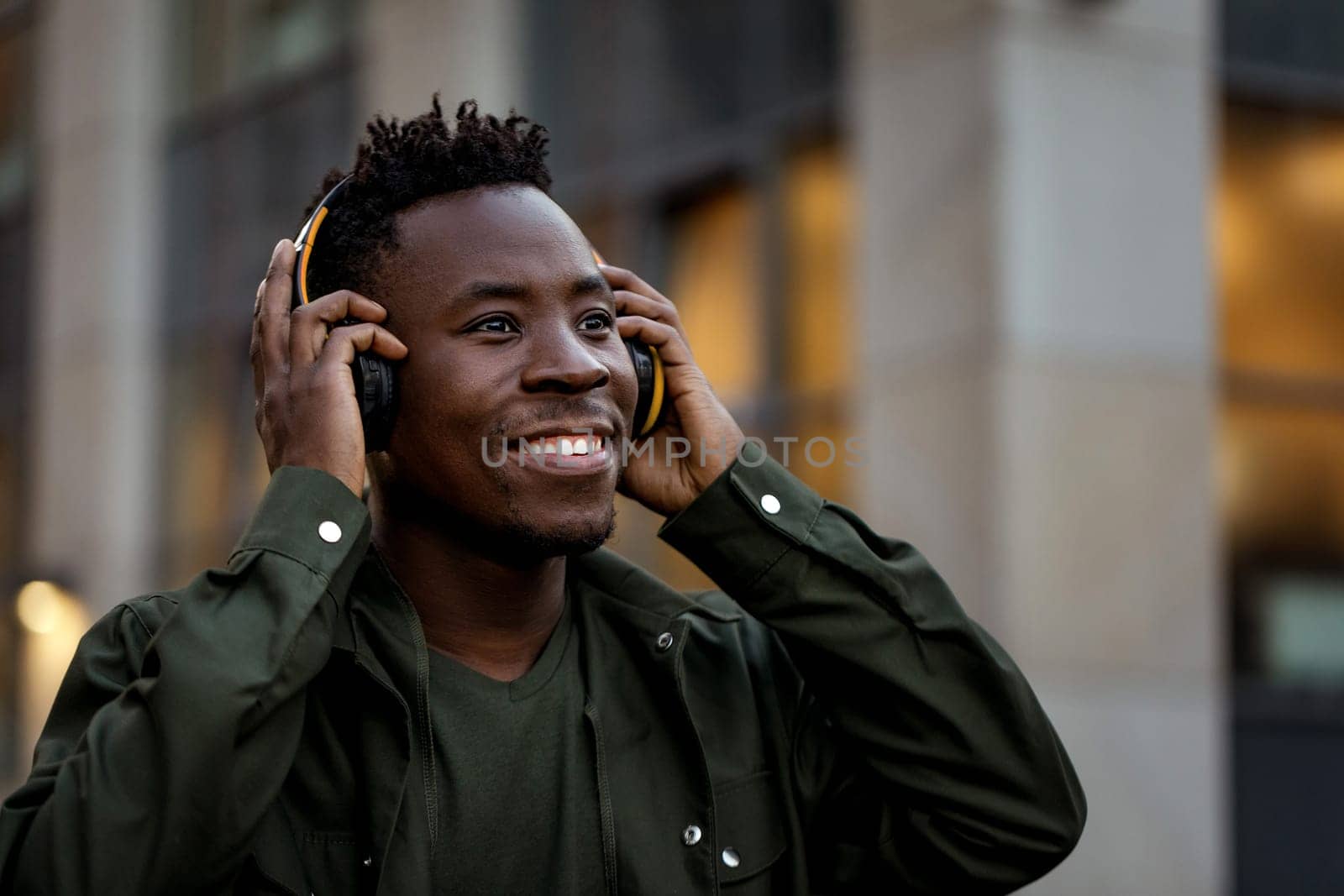 happy african-american man in stylish jacket in wireless headphones listening music on the street of the evening city. space for text