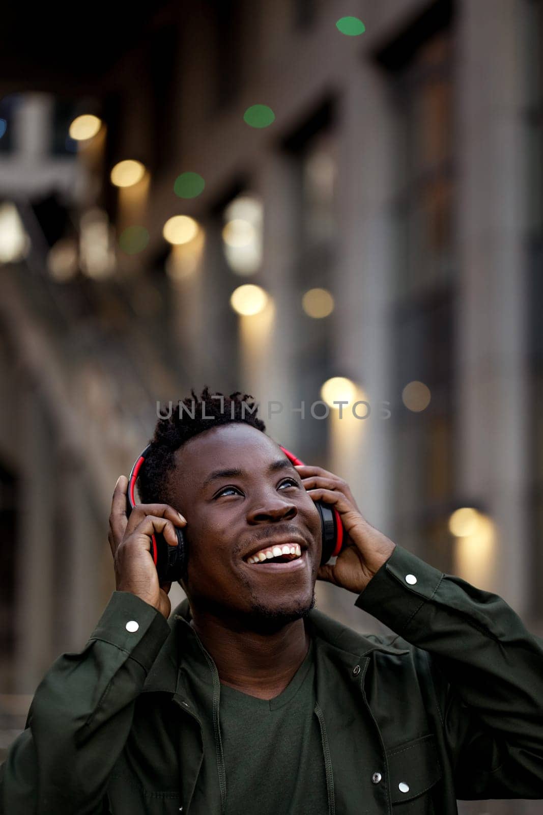 happy african-american man in stylish jacket in wireless headphones listening music on the street of the evening city