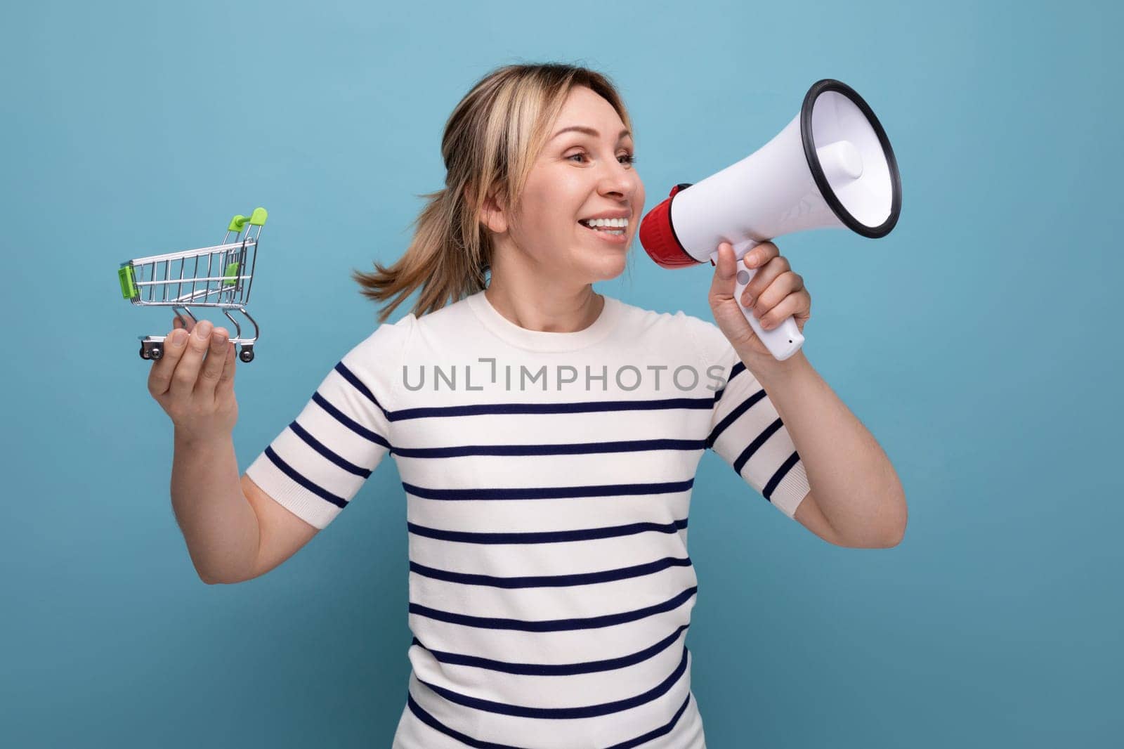 energetic blond attractive young woman in casual outfit announces sale using loudspeaker holding grocery cart on blue background.