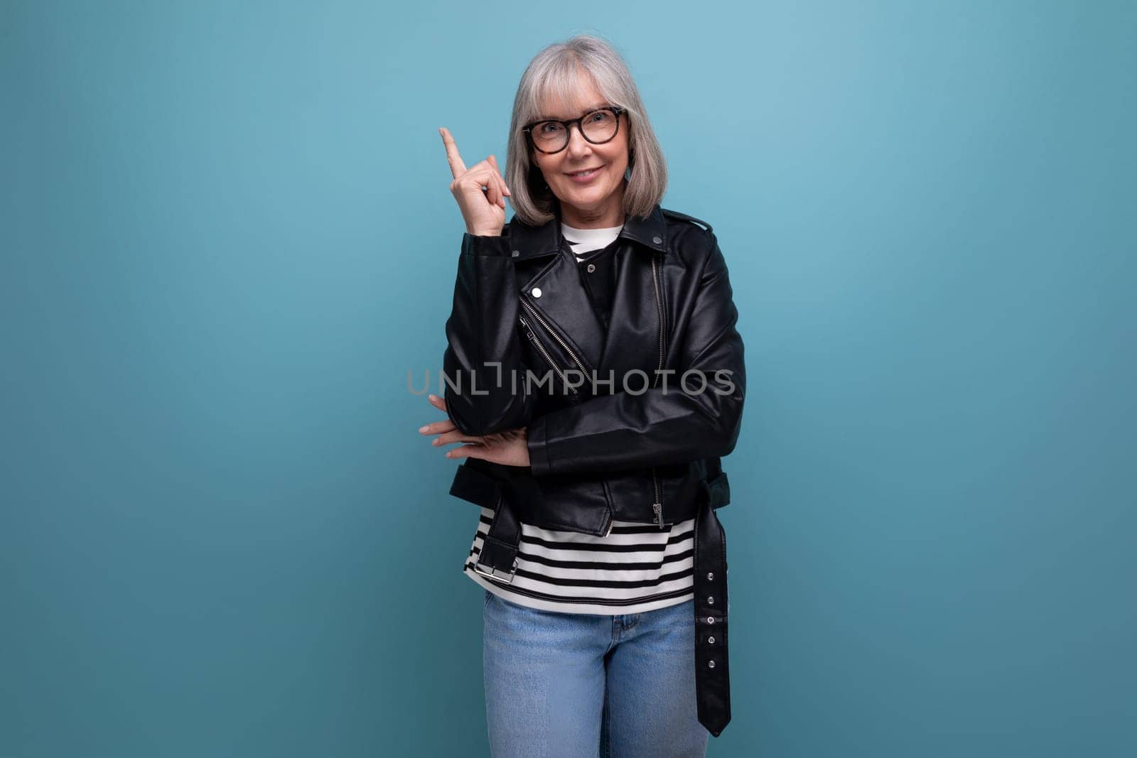 middle-aged pensioner woman in gray hair in a stylish youth look points her finger to the side on a blue background.