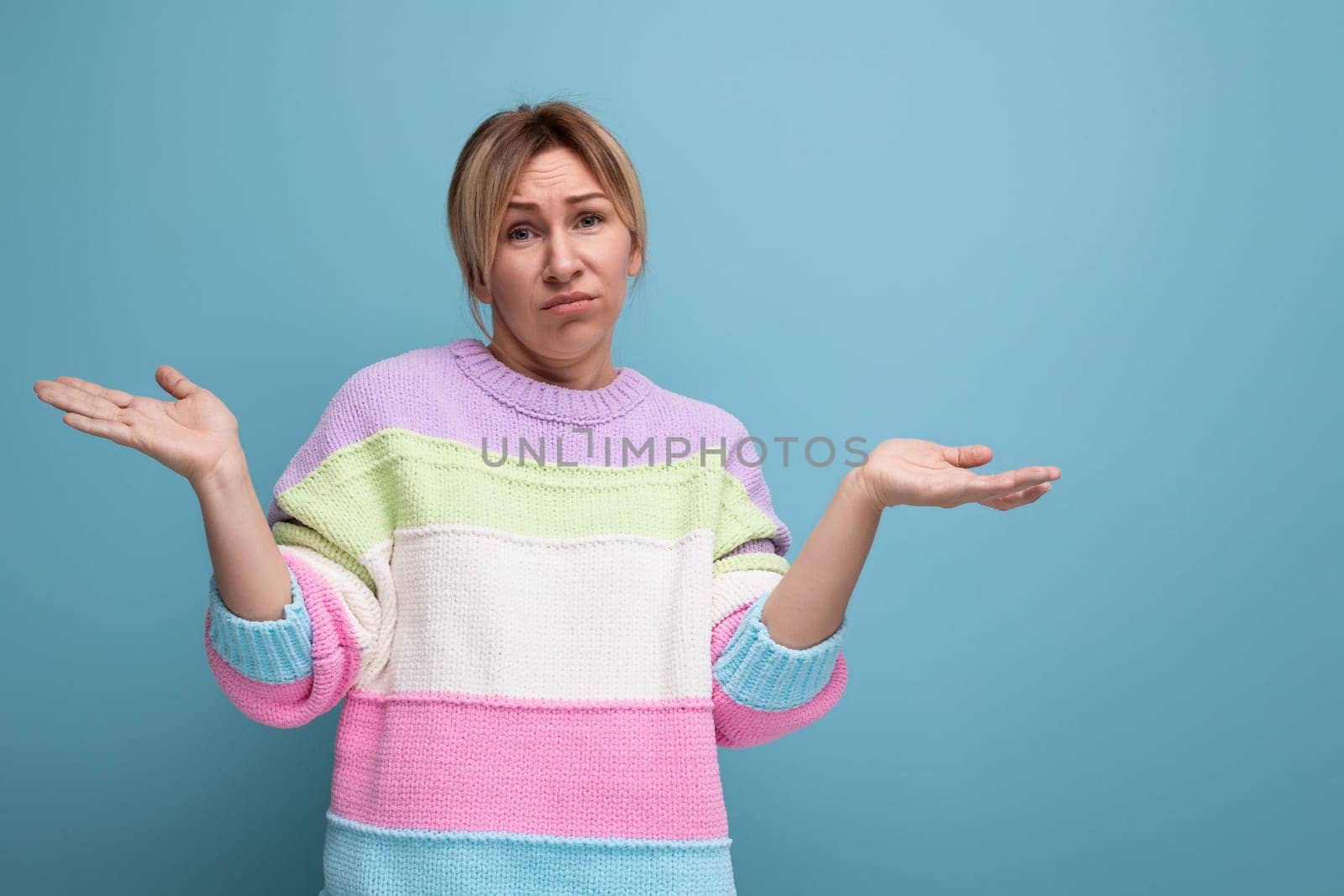 doubtful blond woman in a casual look throws up her hands on a blue background.