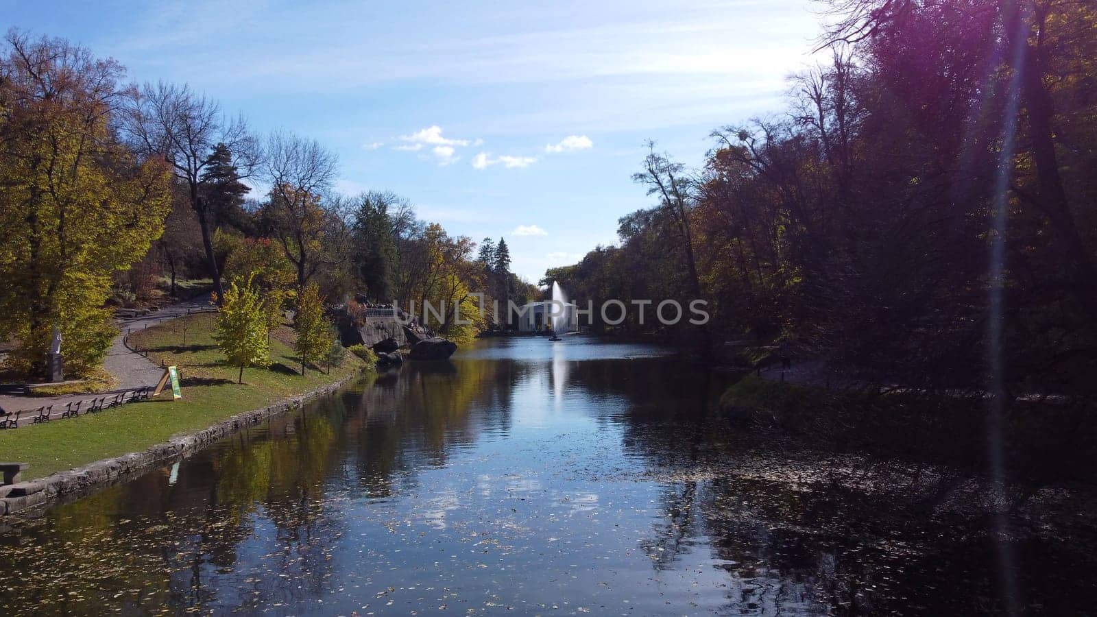 Beautiful landscaped park with decorative fountain in center of lake, trees and large stones on banks on sunny autumn day. Flying over water with fallen leaves and blue sky reflection. Many tourists