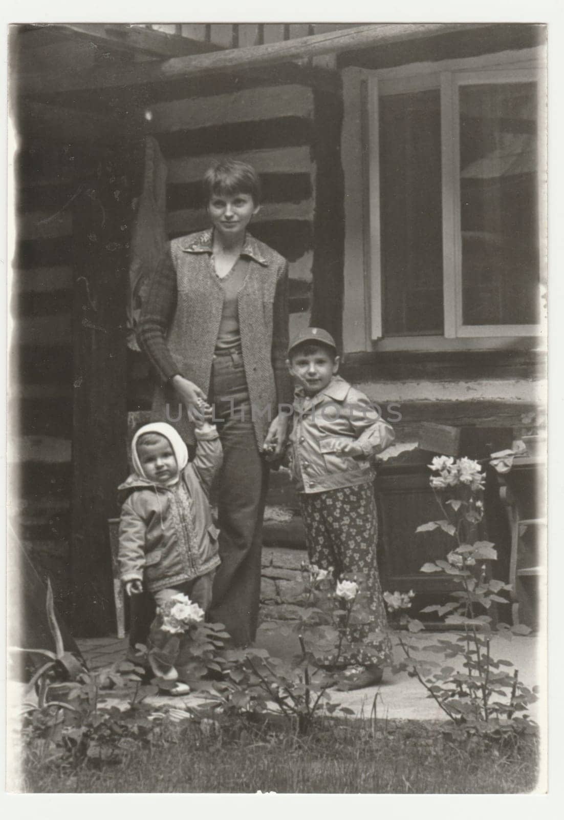 Vintage photo shows an adolescent girl with two children. They pose in front of log cabin. Retro black and white photography. Circa 1980s. by roman_nerud