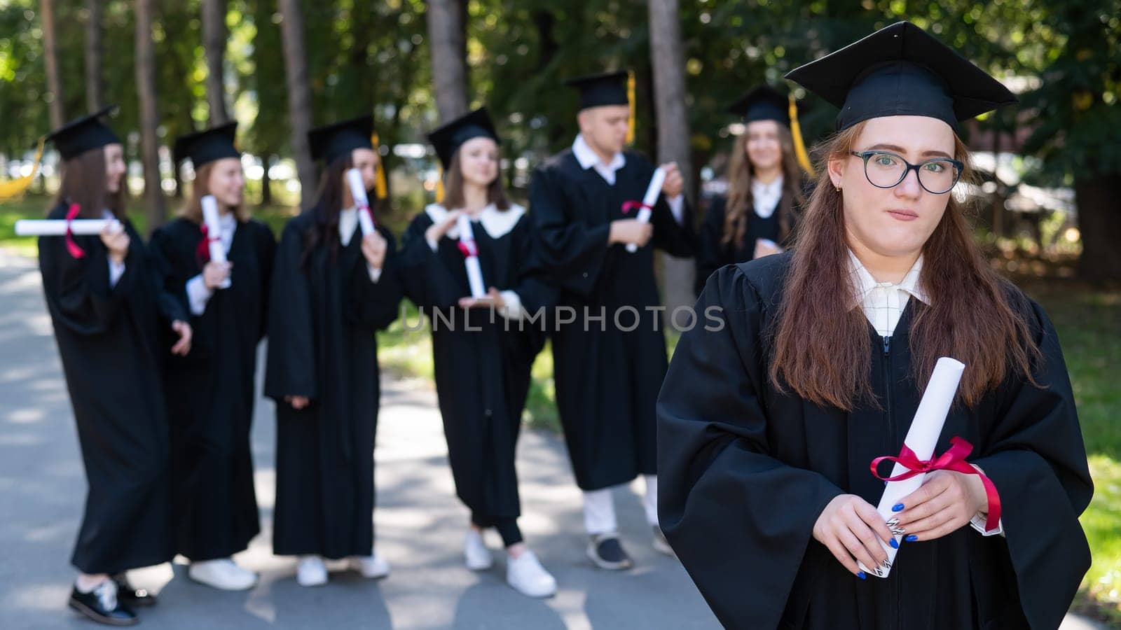 Portrait of a young caucasian woman in glasses and a graduate gown against the background of classmates. A group of graduate students outdoors